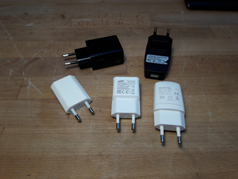 Fünf verschiedene Handynetzteile liegen auf einer Tischplatte.