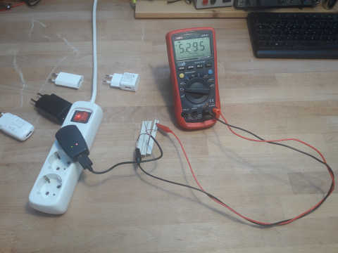 Ein Handynetzteil ist in die Steckdose eingesteckt. Über ein aufgeschnittenes USB Kabel ist ein Widerstand angeschlossen. Am Widerstand ist mit Krokoklemmen ein Spannungsmessgerät angeschlossen.
